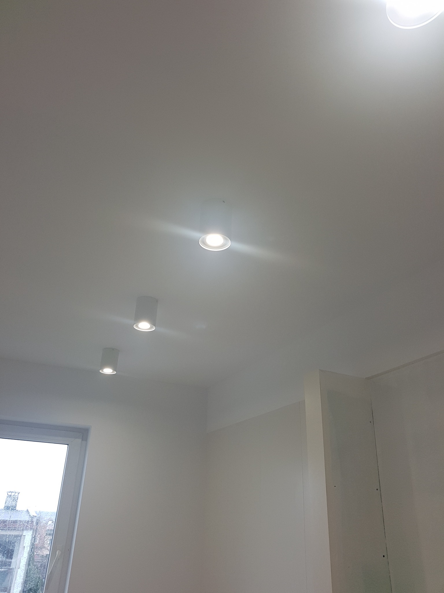 Biały sufit z trzema okrągłymi lampami punktowymi LED, widok z dołu, ściana z płyt kartonowo-gipsowych, okno z widokiem na budynek z cegły.