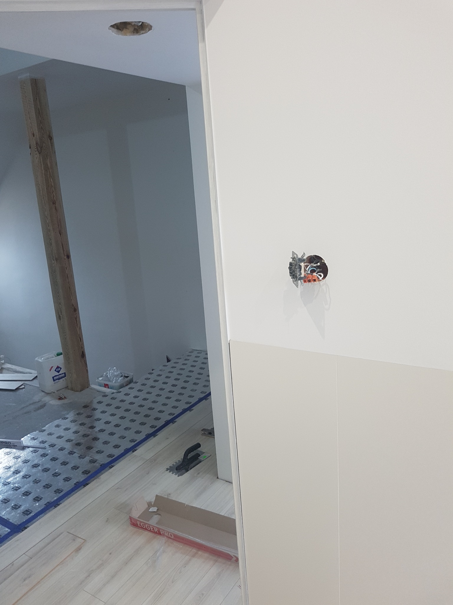 Wnętrze w trakcie prac wykończeniowych: położona podłoga z paneli, widoczna puszka elektryczna bez osprzętu na gładkiej, białej ścianie, oraz drewniany słup konstrukcyjny w tle.