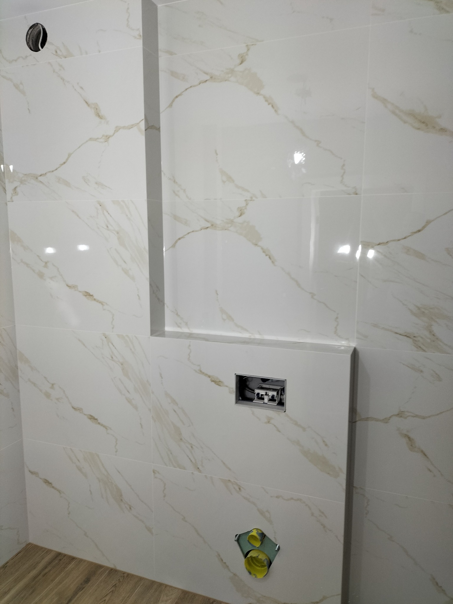 Ściana w łazience wyłożona białymi płytkami imitującymi marmur z widoczną instalacją podtynkową do WC oraz otworem na rurę i fragmentem drewnopodobnej podłogi