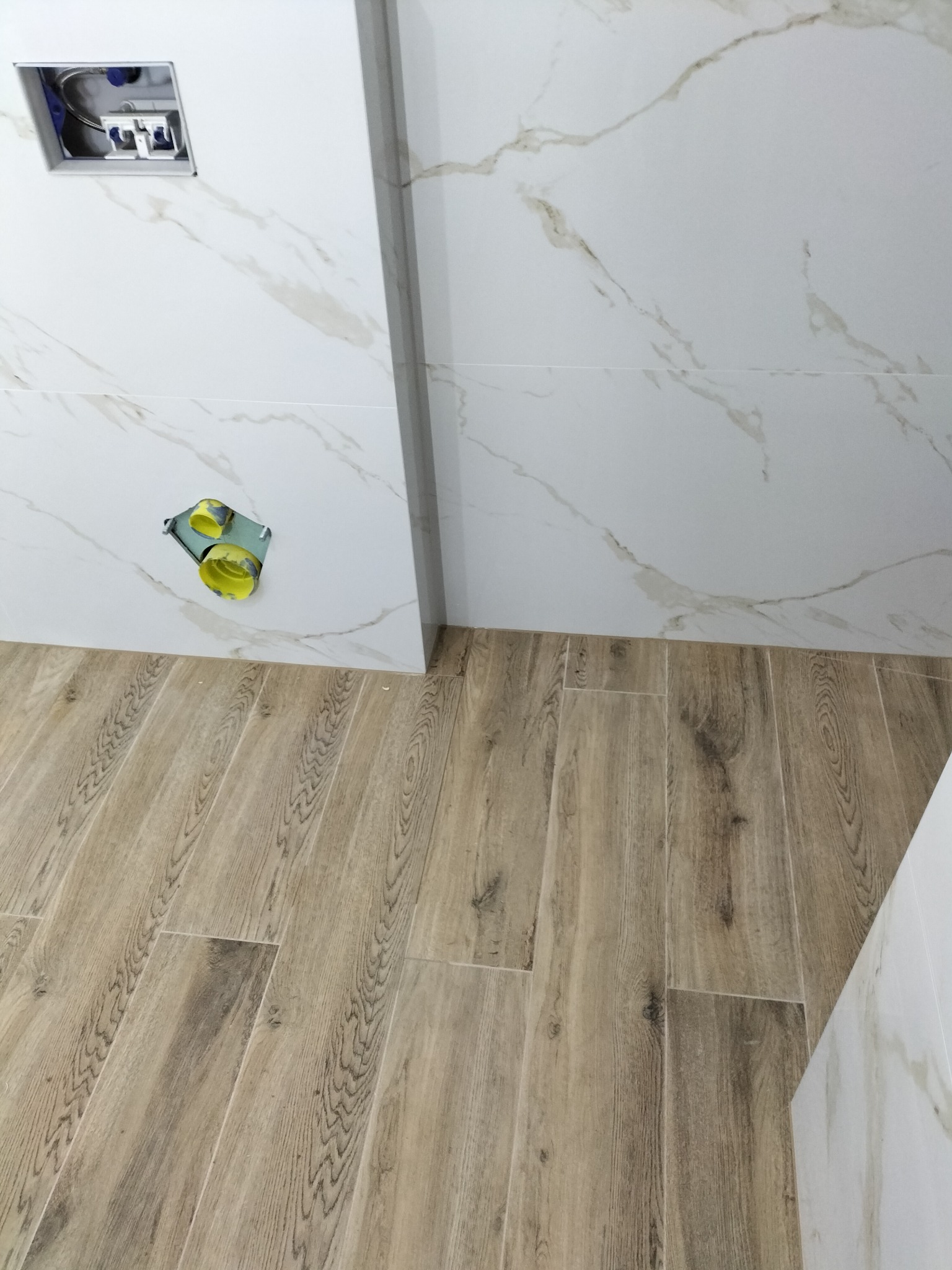 Montaż instalacji podtynkowej z żółtymi elementami w łazience z marmurowymi płytkami ściennymi i podłogą imitującą drewno.