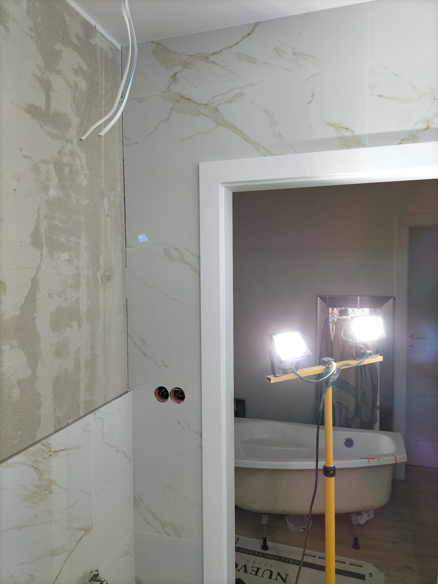 Łazienka w trakcie prac wykończeniowych: częściowo położone marmurowe płytki, fragment ściany z surowym tynkiem, widoczne przewody elektryczne, lampa budowlana oświetlająca wannę w głębi...