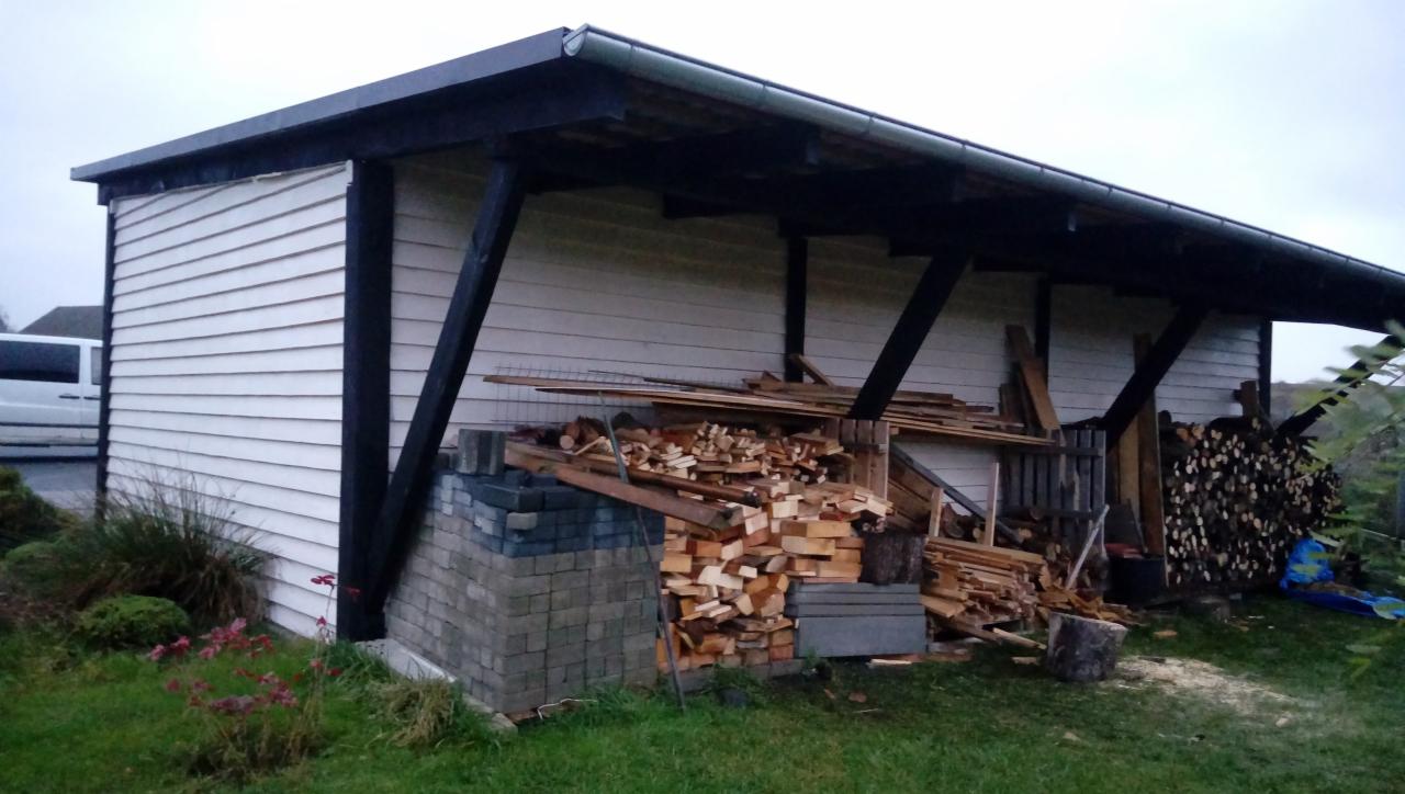 Drewniany garaż z białą elewacją i czarnym dachem, podparty na czarnych, ukośnych belkach; przed garażem stos drewna opałowego ułożony na fundamencie z szarych cegieł.