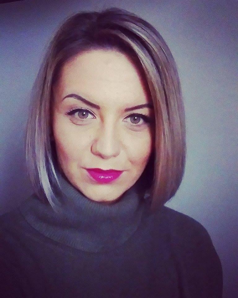 Portret kobiety z blond bobem, mocnym makijażem ust i zielonym golfem na tle szarej ściany.