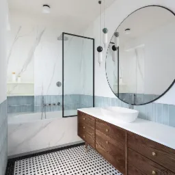 Nowoczesna łazienka z marmurowymi ścianami, błękitnymi akcentami w postaci płytek, czarno-białą mozaiką na podłodze, drewnianą szafką pod umywalkę i okrągłym lustrem.