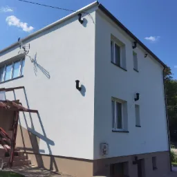 Dwupiętrowy dom z nową, białą elewacją, ciemnymi rynnami i brązowym cokołem, widziany w słoneczny dzień z boku, z widocznym zadaszeniem nad schodami wejściowymi i anteną na dachu.