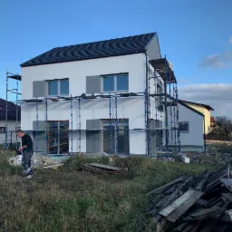 Dwukondygnacyjny dom jednorodzinny w trakcie wykańczania elewacji, z rusztowaniami i pracownikiem w ubrudzonych farbą spodniach, stojącym na tle wysokiej trawy i desek.