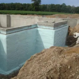 Fundament budynku w trakcie izolacji pianką, widoczny pracownik w kombinezonie ochronnym aplikujący piankę na ściany fundamentu, w tle zielone pole i drzewa.