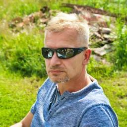 Portret mężczyzny w okularach przeciwsłonecznych z odbiciem grupy osób przy stole w soczewkach, na tle zielonej trawy i zarośli.