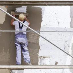 Pracownik na rusztowaniu nakłada szarą zaprawę na ścianę budynku ocieplonego styropianem, widoczny fragment okna.