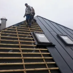 Montaż blachy na dachu z oknami dachowymi; widoczna konstrukcja drewniana i pracownicy na górze.