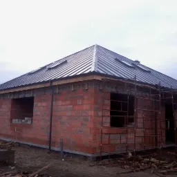 Niedokończony budynek z czerwonej cegły z nowym, ciemnym, metalowym dachem z oknami dachowymi, częściowo osłonięty rusztowaniem.