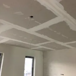 Sufit z płyt kartonowo-gipsowych w trakcie szpachlowania łączeń, widoczne otwory na oświetlenie i wystające kable, ściany częściowo pomalowane na szaro, okno z butelką wody na parapecie.