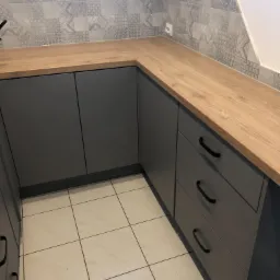 Szare szafki kuchenne z drewnianym blatem i czarnymi uchwytami szuflad, nad nimi dekoracyjne płytki ścienne w odcieniach szarości i beżu, a pod nimi jasne płytki podłogowe.