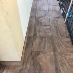 Wyłożona brązowymi płytkami podłoga balkonu, widoczna ściana budynku pomalowana na dwa odcienie beżu oraz metalowa balustrada