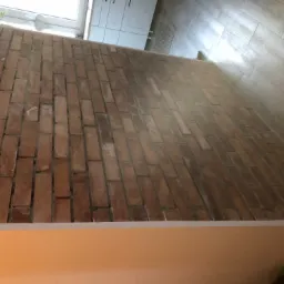 Ściana wyłożona cegłą klinkierową w odcieniach czerwieni i brązu, z widocznymi fugami, sąsiadująca z białymi szafkami i jasną podłogą z płytek imitujących drewno.