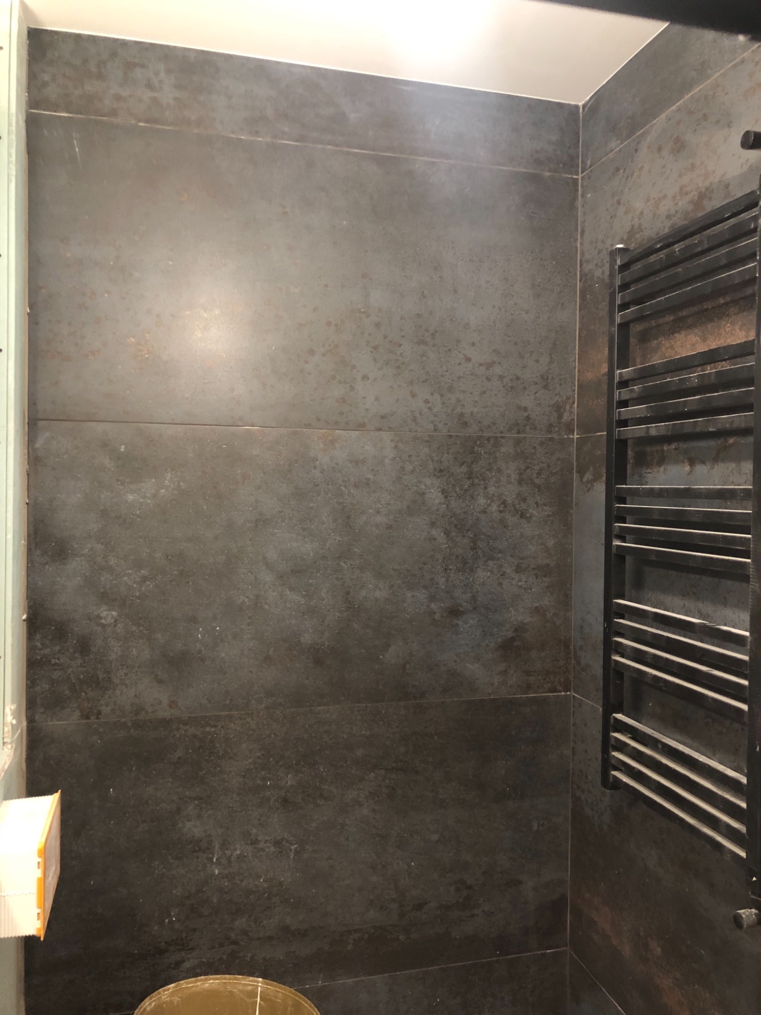 Ciemne, metaliczne płytki w kabinie prysznicowej z grzejnikiem drabinkowym po prawej stronie, widoczny fragment wiadra na dole kadru.