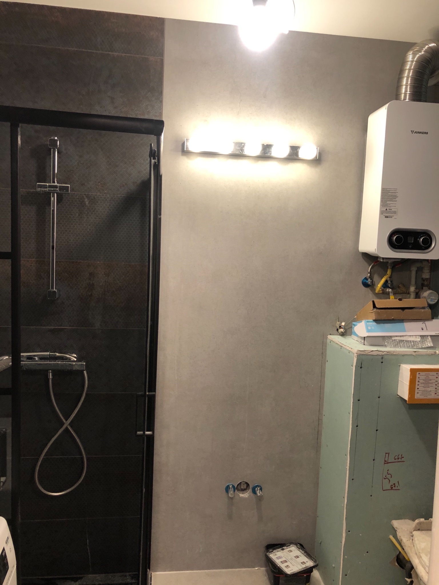 Częściowo wykończona łazienka z ciemnymi płytkami w kabinie prysznicowej, jasnoszarą ścianą, grzejnikiem Junkers i obudową z płyty gipsowo-kartonowej z widocznymi wymiarami.