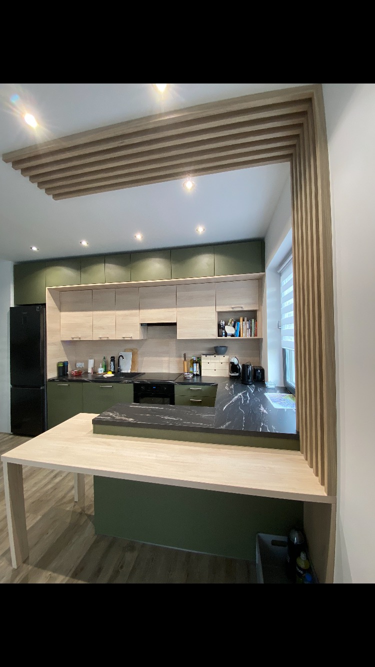 Nowoczesna kuchnia z drewnianymi lamelami na suficie i ścianie, zielonymi i beżowymi szafkami oraz blatem z czarnego marmuru.
