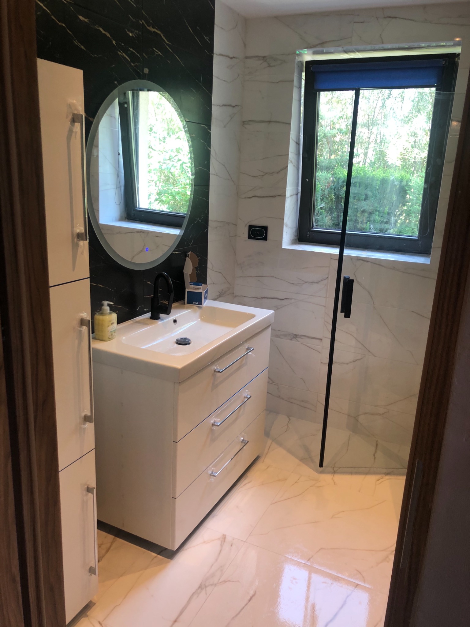 Nowoczesna łazienka z białymi marmurowymi płytkami, owalnym lustrem z podświetleniem LED, białą szafką z umywalką i czarną armaturą, oraz kabiną prysznicową ze szklanymi drzwiami z czarnymi detalami.