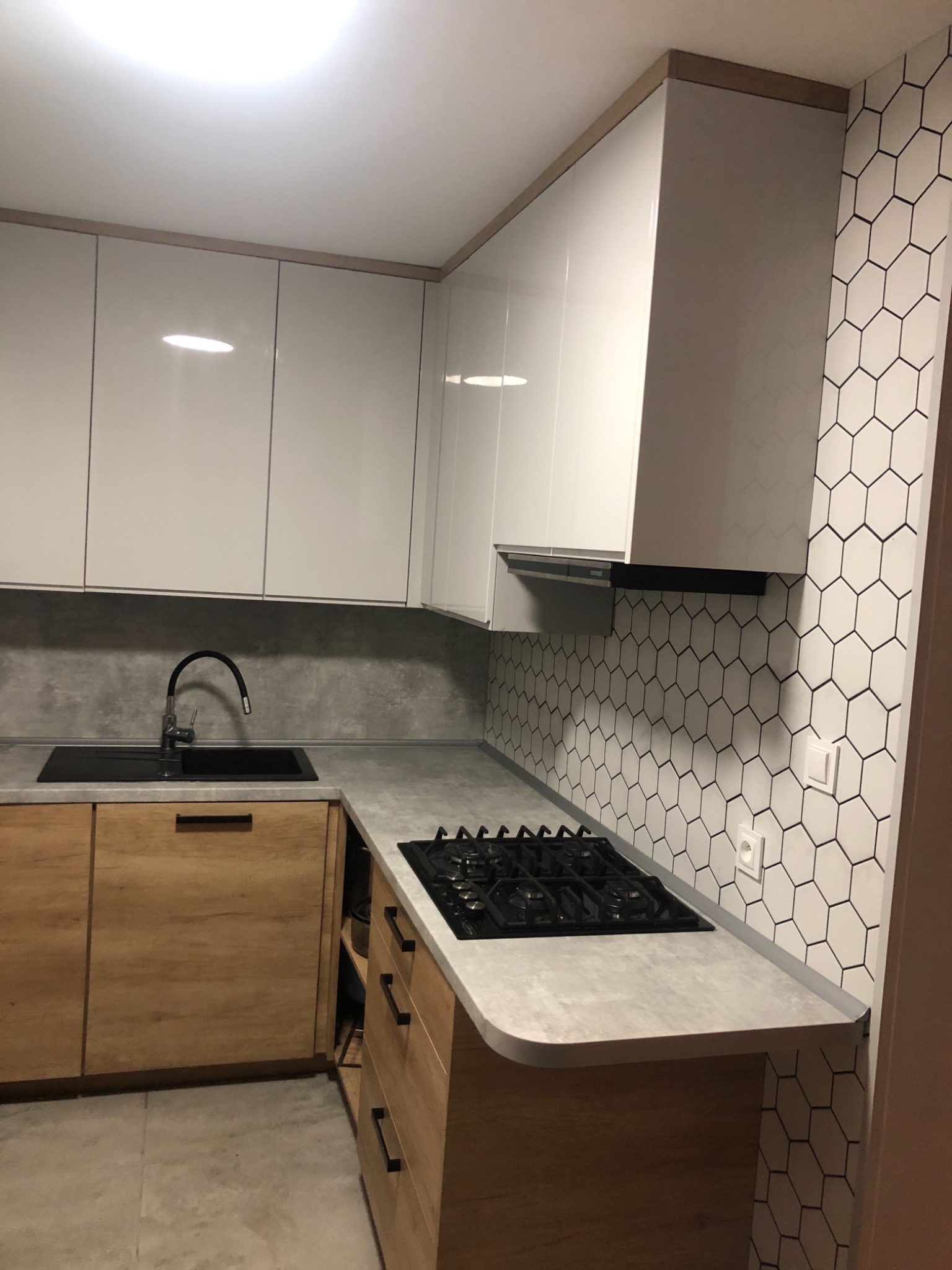 Nowoczesna kuchnia z białymi, lakierowanymi szafkami górnymi, drewnianymi szafkami dolnymi z czarnymi uchwytami, czarnym zlewem i baterią oraz płytą grzewczą, wykończona heksagonalną mozaiką...