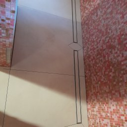Bud Emp - Beżowe płytki podłogowe z nowoczesnym liniowym odpływem o geometrycznym wzorze. Ściana wyłożona drobną mozaiką w odcieniach różu i czerwieni.