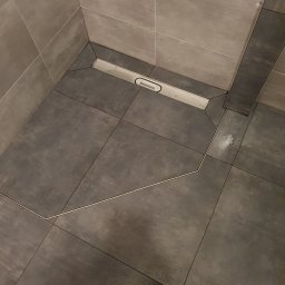 Bud Emp - Precyzyjnie ułożone ciemnoszare płytki podłogowe w kabinie prysznicowej, zintegrowane z liniowym odpływem. Jasnoszare płytki ścienne i metalowe listwy tworzą nowoczesny design.