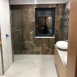 Nowoczesna łazienka z kabiną prysznicową typu walk-in, wykończona płytkami imitującymi kamień w odcieniach brązu i beżu, z dużym oknem i grzejnikiem drabinkowym.