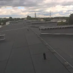 Płaski dach pokryty czarną papą termozgrzewalną z widocznymi kominkami wentylacyjnymi i elementami obróbek blacharskich, w tle zabudowa miejska i pochmurne niebo.