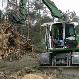 Zielono-biała koparka kołowa z chwytakiem podnosi wykopany korzeń drzewa na tle stosów pociętego drewna i lasu.