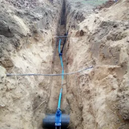 Wykopany wąski rów w ziemi z widocznymi niebieskimi rurami wodociągowymi i czarną rurą kanalizacyjną w trakcie instalacji, na powierzchni widoczny pracownik i budynek w trakcie budowy.