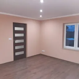 Pomalowane na jasny r&oacute;ż pok&oacute;j z brązową podłogą, ciemnymi drzwiami z poziomymi szybami, białym oknem i białymi listwami sufitowymi, oświetlony trzema punktowymi lampami sufitowymi.