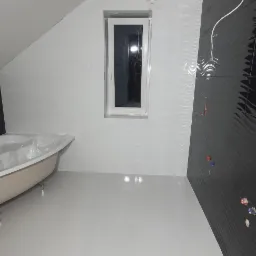 Nowoczesna łazienka na poddaszu z białą podłogą i ścianami wykończonymi płytkami oraz czarną ścianą z dekoracyjnymi falami. Wanna w folii ochronnej stoi pod oknem. Widoczne niedokończone instalacje...