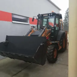 Pomarańczowo-czarna koparko-ładowarka Case z dużą czarną łyżką z przodu, zaparkowana na betonowym placu przed szaro-białym budynkiem z poziomym czerwonym pasem.