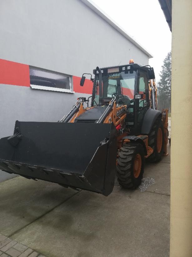 Pomarańczowo-czarna koparko-ładowarka Case z dużą czarną łyżką z przodu, zaparkowana na betonowym placu przed szaro-białym budynkiem z poziomym czerwonym pasem.