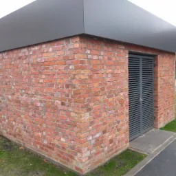 Niewielki budynek z cegły rozbiórkowej z szarą, metalową kratą w roli drzwi i płaskim dachem, otoczony trawnikiem i chodnikiem.