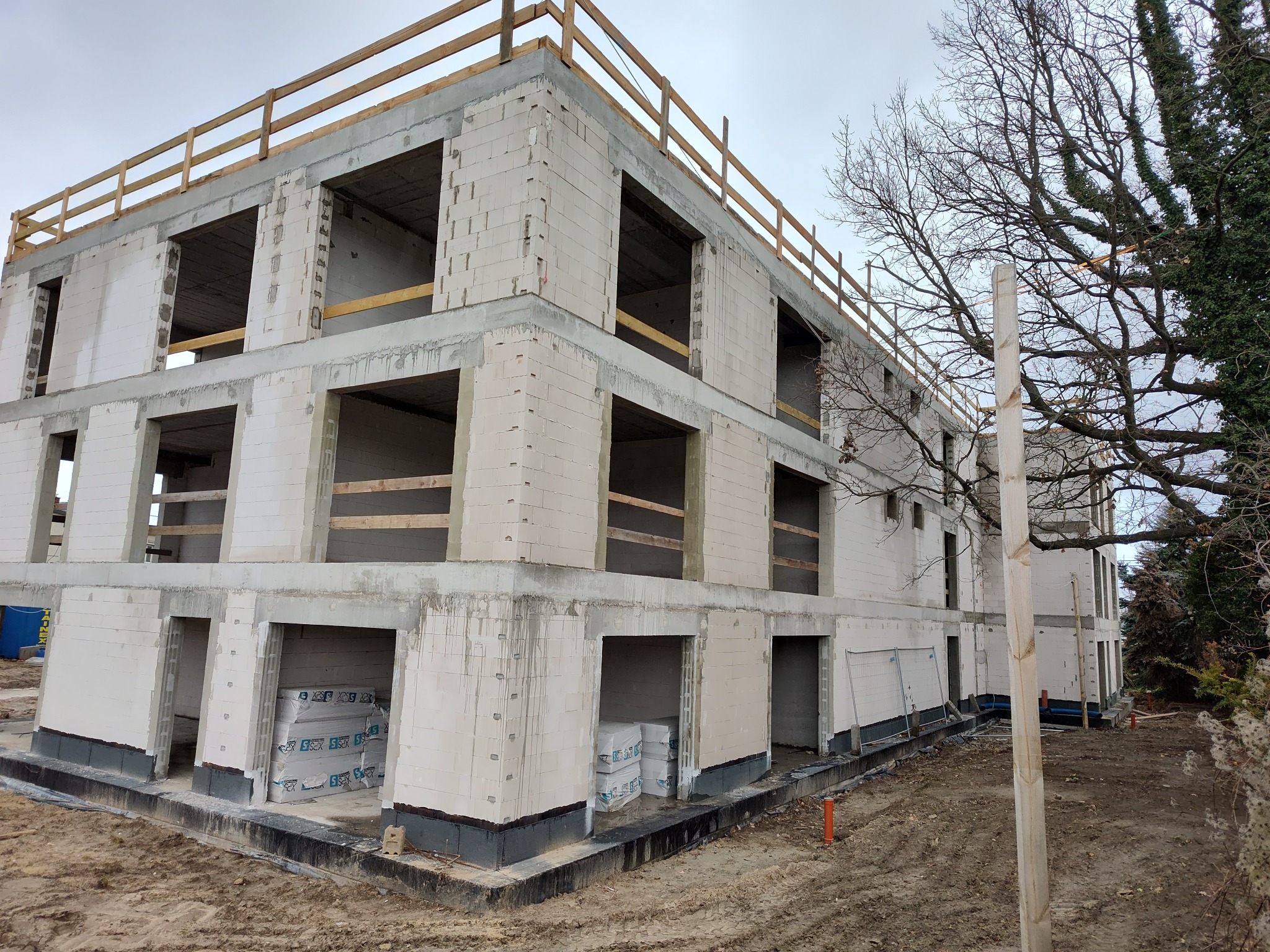 Wznoszenie dwukondygnacyjnego budynku biurowego z białych bloczków, widoczne otwarte przestrzenie okienne i drzwiowe, zabezpieczone drewnianą barierką na dachu, na pierwszym planie ułożone paczki...