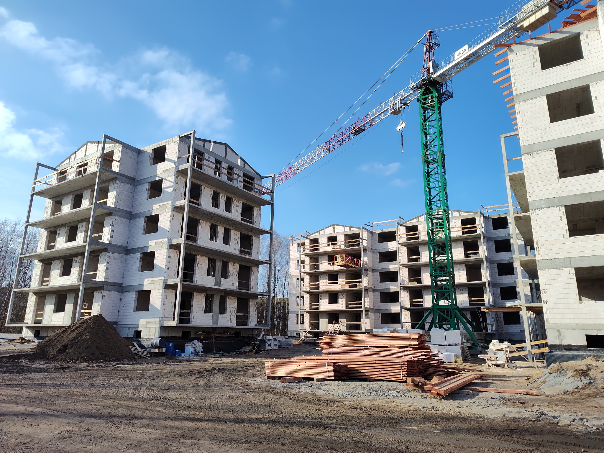 Budowa stanu surowego otwartego trzech budynków mieszkalnych wielorodzinnych z betonu komórkowego, widoczny żuraw wieżowy, stos drewna budowlanego i hałda ziemi.