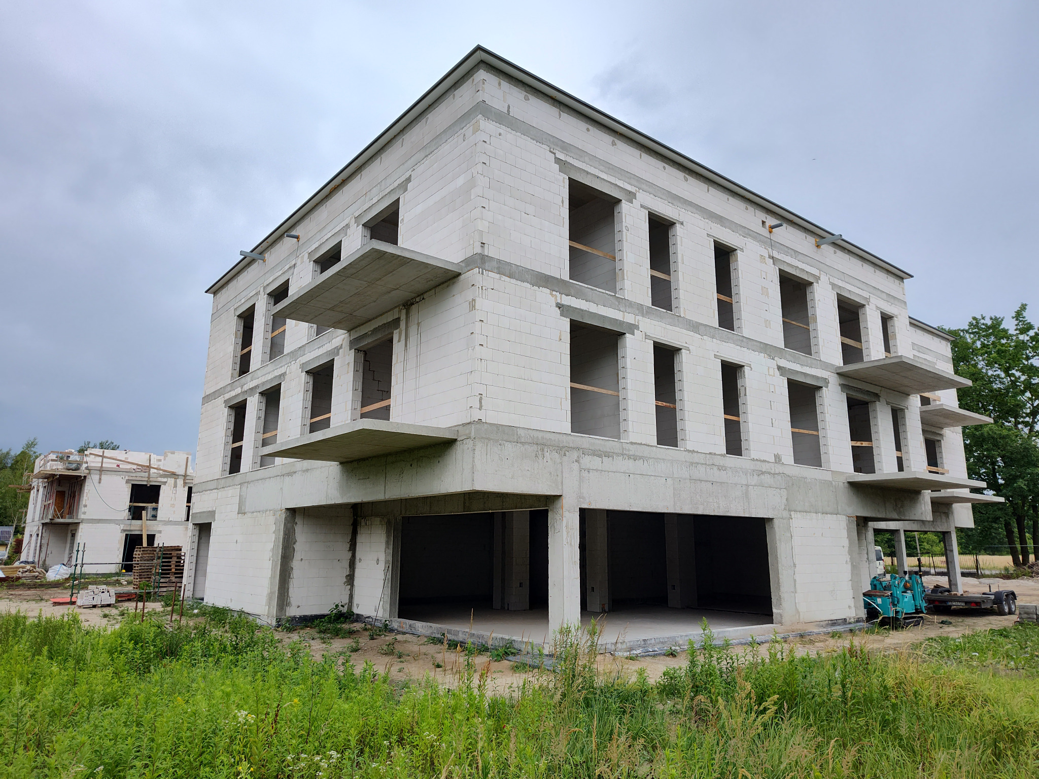 Budynek wielorodzinny w stanie surowym otwartym, z betonu komórkowego, z widocznymi elementami żelbetowymi, na tle pochmurnego nieba i zieleni. Widoczny sprzęt budowlany.
