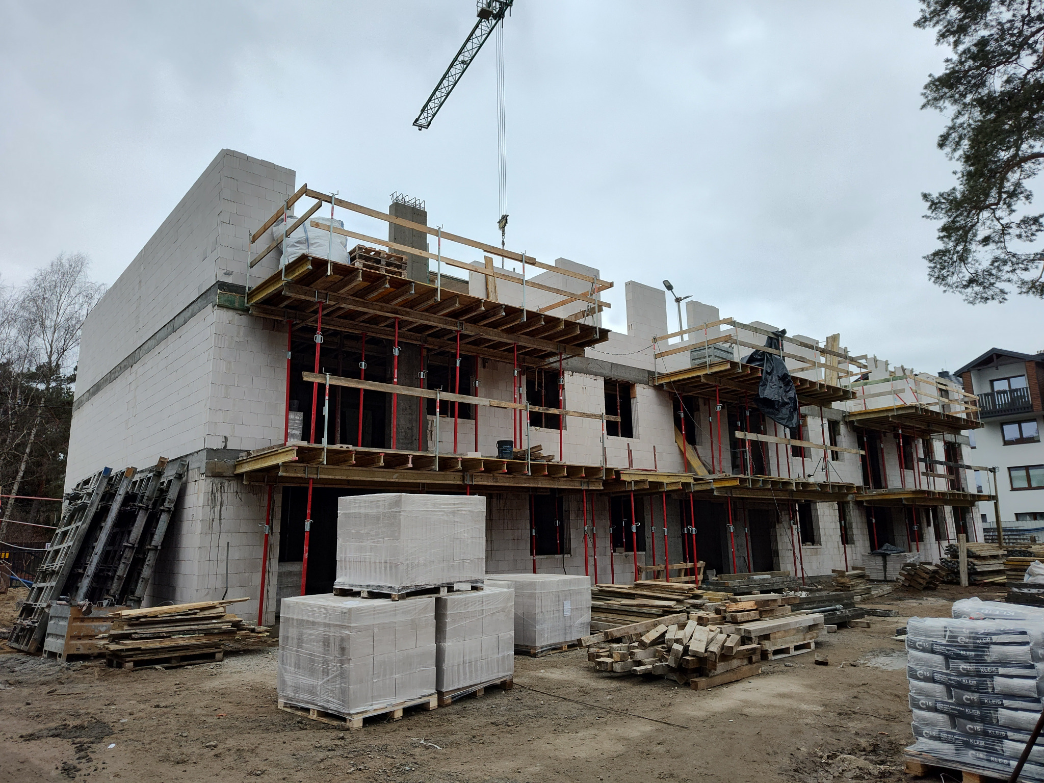 Budowa stanu surowego otwartego: dwukondygnacyjny budynek wielorodzinny z białego bloczka, widoczne rusztowania, dźwig budowlany i materiały budowlane.