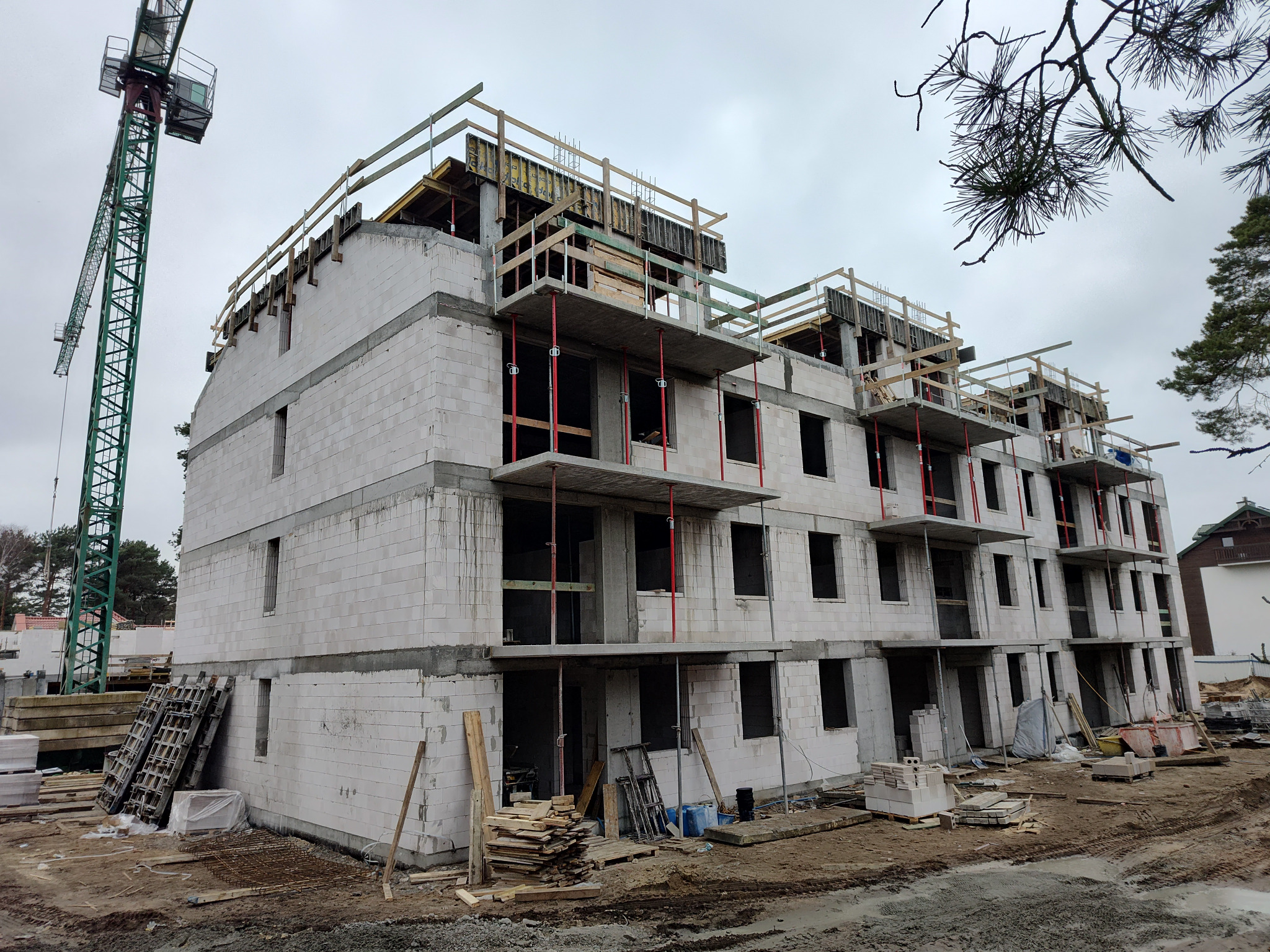 Budowa stanu surowego otwartego: wielopiętrowy budynek z białych bloczków, widoczne rusztowania, dźwig budowlany w tle, teren budowy.