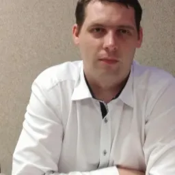 Portret mężczyzny w białej koszuli z podwiniętymi rękawami na tle beżowej ściany. Zdjęcie profilowe 'Głażewski'.