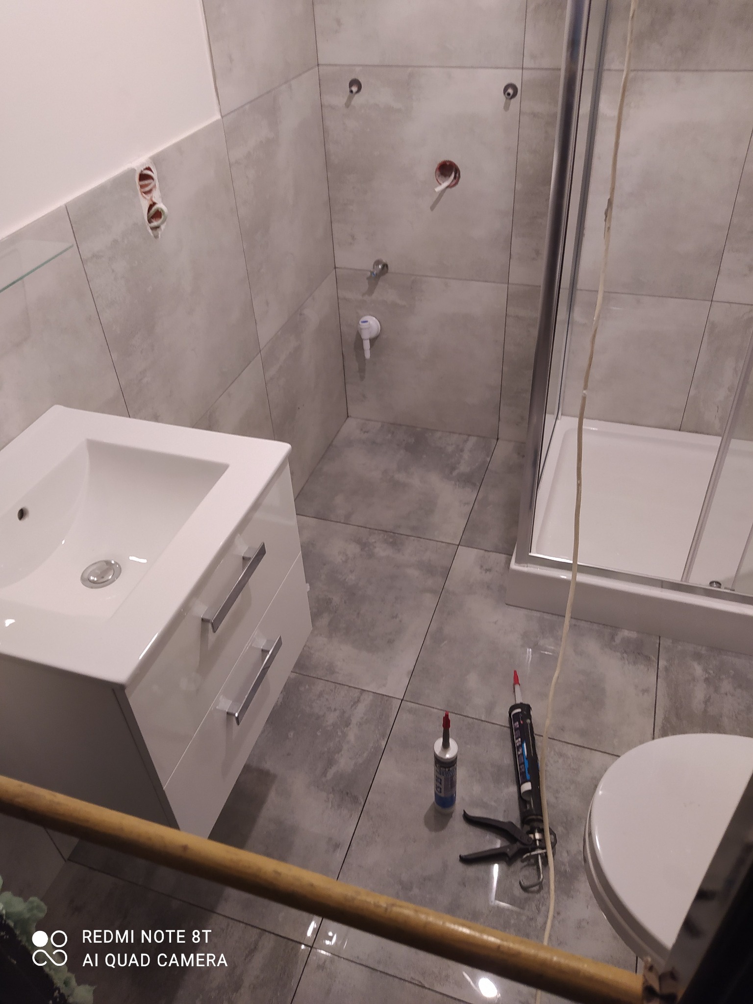 Wnętrze łazienki w trakcie wykańczania, widoczna umywalka z szafką, kabina prysznicowa, szare płytki na ścianach i podłodze, oraz narzędzia i materiały do fugowania i silikonowania.