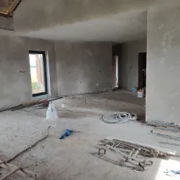 Wnętrze w stanie surowym po nałożeniu tynków, widoczne przewody elektryczne wystające ze ścian, resztki materiałów budowlanych i narzędzia na podłodze.