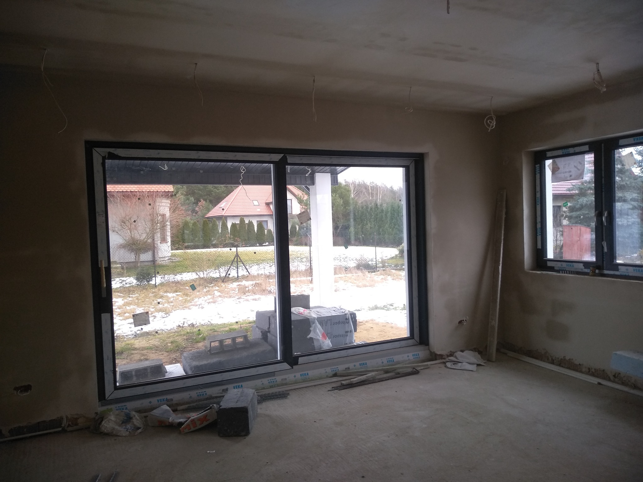 Surowy stan ścian w Dłutowie po nałożeniu tynku, widoczne wystające kable, duże okno przesuwne z widokiem na ośnieżony ogród i nieotynkowane ściany przy podłodze.