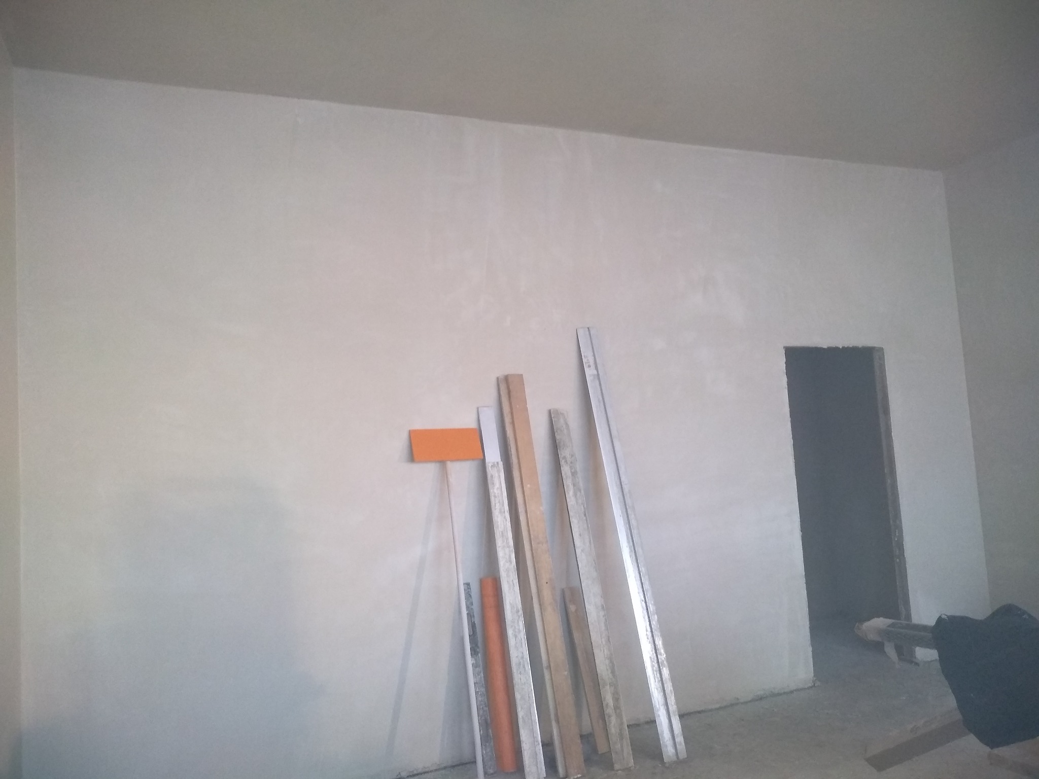 Świeżo otynkowana ściana w pokoju z widocznymi narzędziami i łatami po lewej stronie oraz otworem drzwiowym w tle.