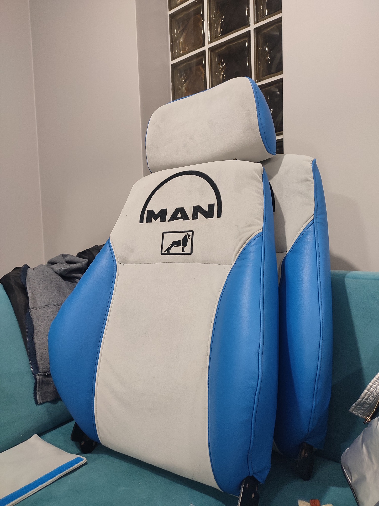 Odnowione siedzenie kierowcy MAN z biało-niebieską tapicerką, widoczne logo firmy, stojące na turkusowej kanapie.