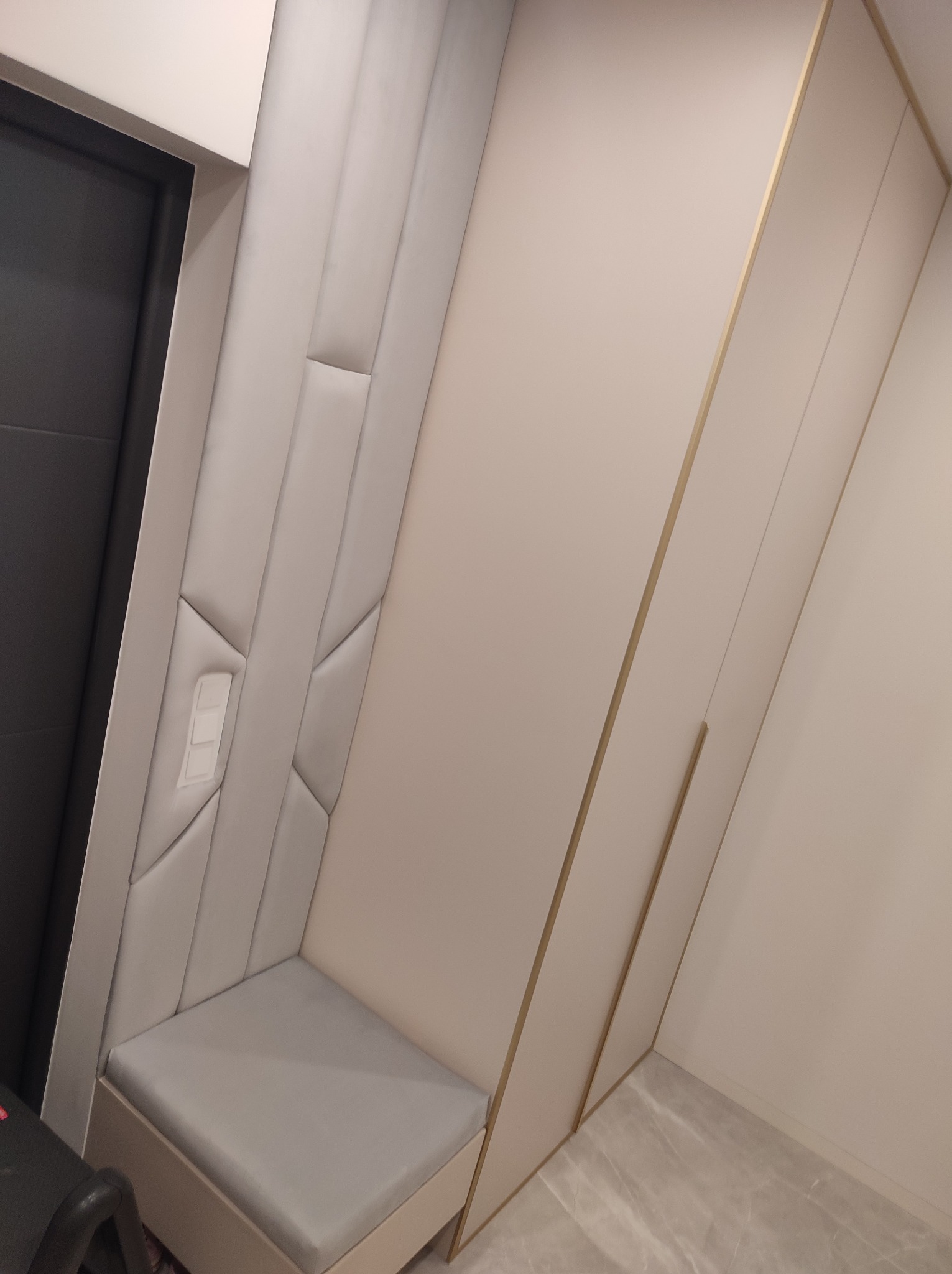 Tapicerowany panel ścienny z geometrycznym wzorem i wbudowanym siedziskiem w przedpokoju, obok szafy wnękowej z subtelnymi złotymi akcentami.