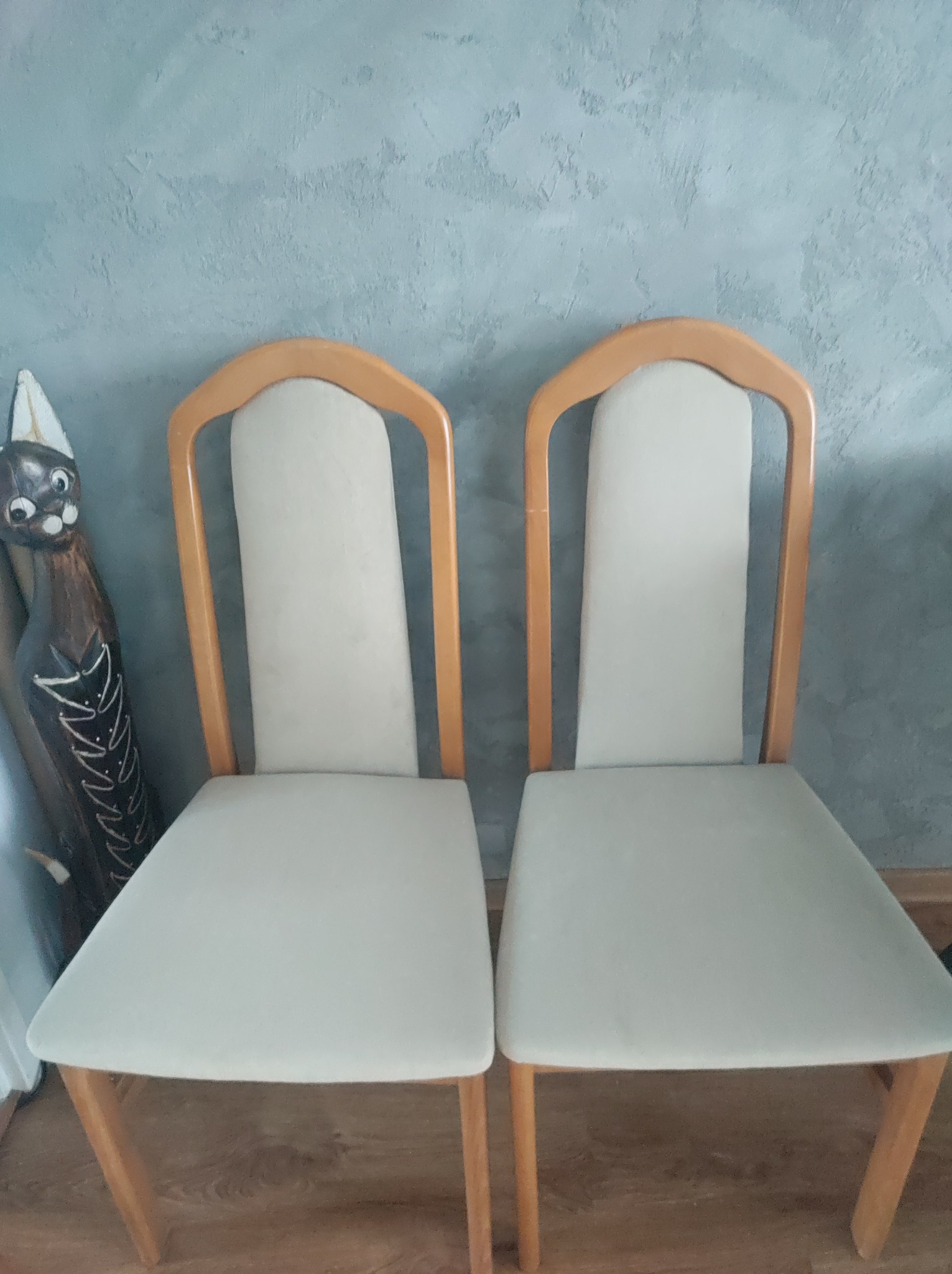 Dwa krzesła z jasną tapicerką i drewnianymi ramami stoją obok drewnianej figurki kota na tle szarej ściany.