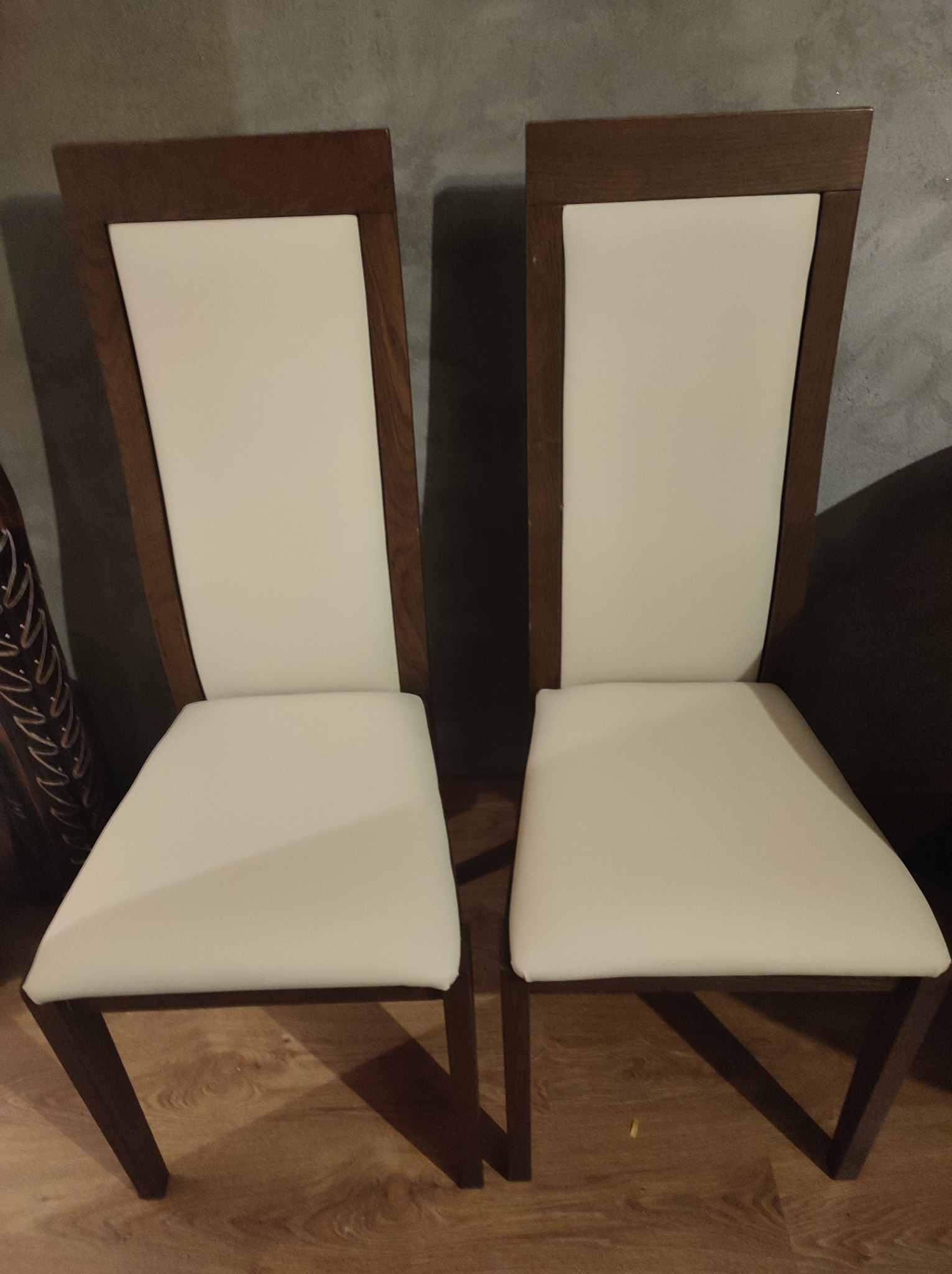 Dwa krzesła z ciemnego drewna i jasną tapicerką, widoczne z przodu, ustawione na drewnianej podłodze.