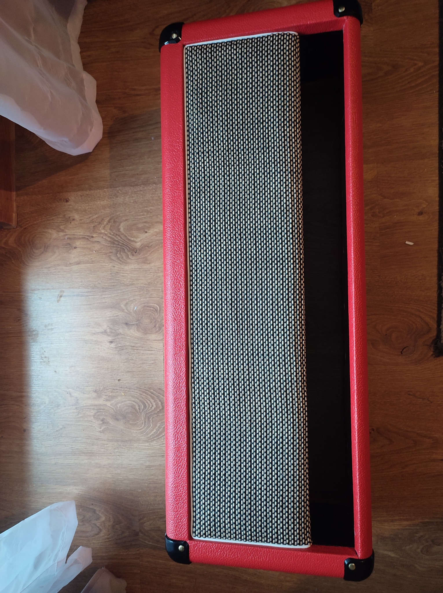 Czerwona obudowa wzmacniacza gitarowego z czarno-białą maskownicą, widoczna na drewnianej podłodze, z fragmentem białej folii w lewym górnym rogu.